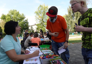 Chłopcy stoją przy stoliku, gdzie leżą kartki z kamienicami do malowania i pudełka z kredkami.