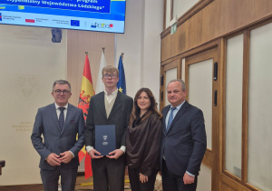 Nagrodzony uczeń w otoczeniu nauczycielki i wręczających listy gratulacyjne.