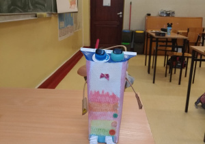 Robot uczennicy wykonany z kartonu po mleku, ozdobiony nakrętkami, guzikami i doklejonym, pomalowanym papierem z pismem brajla