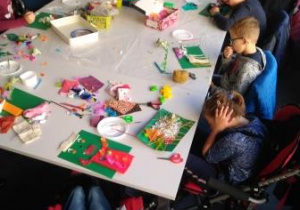 Uczniowie siedzący przy stole wykonują własne stwory z różnorodnych materiałów: kolorowych kartek, bibuły, guzików, kolorowych piórek itp.