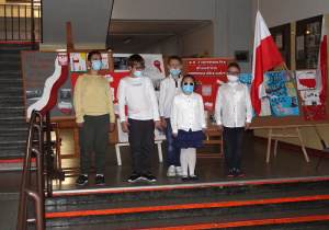 Uczniowie klas czwartych ubrani galowo śpiewają hymn Polski. Za ich plecami prace plastyczne związane ze Świętem Niepodległości. Po prawej stronie białoczerwone flagi.