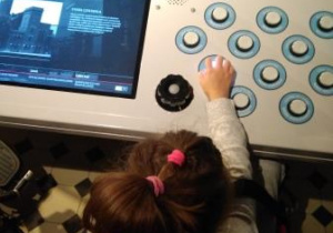 Dziewczynka naciska jeden z przycisków na panelu sterowniczym. Po naciśnięciu przycisk świeci. Po lewej stronie widoczny jest monitor z wyświetlającą się prezentacją o EC1. Na wprost dziewczynki – kolorowa makieta EC1 w przeszklonej witrynie.