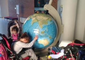Trzy uczennice zgromadzone są wokół dużego globusa zamontowanego na uchwycie przy wysokim słupie. Jedna z uczennic trzyma rękę na globusie i próbuje wprawić go w ruch.