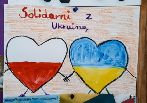 Rysunek dwóch serc trzymających się za ręce, serca wypełnione barwami narodowymi Polski i Ukrainy, napis "Solidarni z Ukrainą".