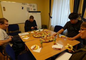 Uczniowie siedzący przy stole. Stoją na nim talerze z kanapkami i owocami.