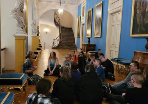 Uczniowie siedzą na podłodze i słuchają przewodnika.