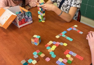 Dziewczynka i chłopiec budują z klocków bryły. Na stole leżą ułożone z klocków litery: M, A, T i E.