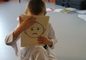 Chłopiec zakrywa twarz rysunkiem smutnej buzi.