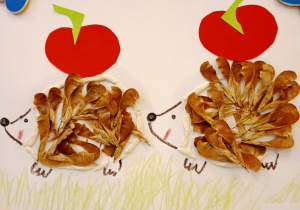 Dwa jeże wykonane z suszonych listków, na grzbietach wycięte papierowe czerwone jabłuszka.