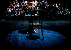 Na scenie stoi krzesło, w tle osoby siedzące na widowni.
