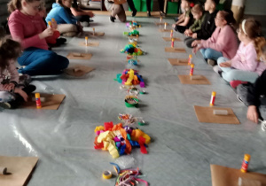 Dzieci siedzą na podłodze. Przed nimi materiały do wykonania prac plastycznych.
