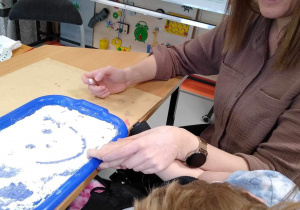 Nauczycielka pokazuje dziewczynce uśmiechniętą buzię narysowaną palcem na rozsypanej mące.