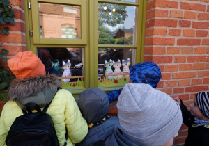 Uczniowie stoją przed oknem budynku, w którym wstawione są figurki kotów.