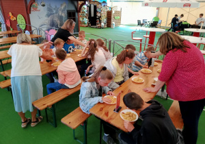 Dzieci siedzą przy stołach i jedzą posiłek.