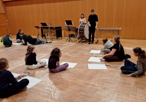Uczniowie siedząc na podłodze słuchają pracownika filharmonii.