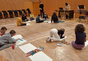 Uczniowie siedząc na podłodze słuchają pracownika filharmonii.