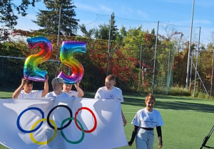 Uczniowie z balonowymi cyframi 2 i 5 oraz flagą olimpijską.