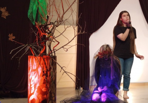 Na scenie stoi drzewo z dużym pniem i gałęziami bez liści. Postać kobiety klęczy przed drzewem z tiulem na głowie. Druga postać stoi za nią z mikrofonem.