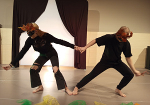 Dwoje ludzi w maskach teatralnych przybranych jesiennymi liśćmi stoi przodem do publiczności. Trzymają się za ręce i wykonują krok - każdy w inną stronę.
