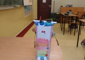 Robot uczennicy wykonany z kartonu po mleku, ozdobiony nakrętkami, guzikami i doklejonym, pomalowanym papierem z pismem brajla